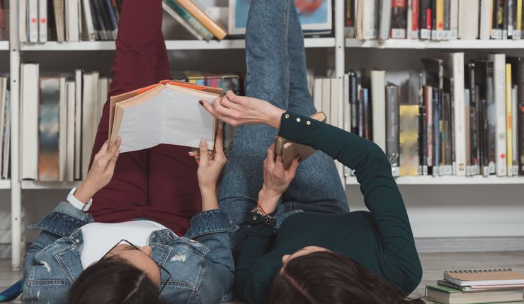 Bücherregal, davor zwei Mädchen lesend auf dem Rücken liegend, die Beine an das Regal gelehnt